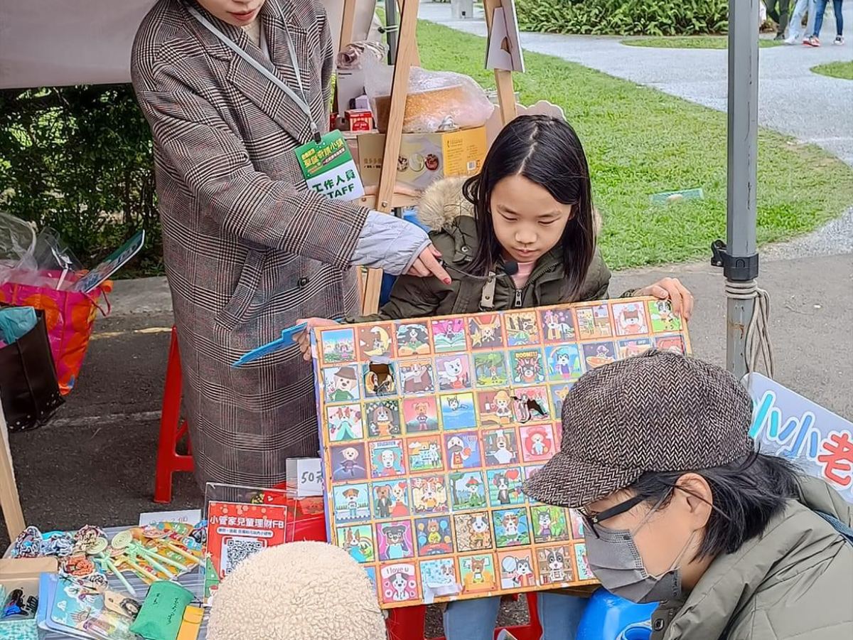 老師陪孩子在小老闆市集攤位上，向客人介紹自己做的商品